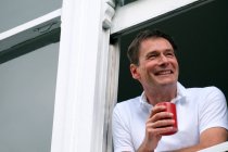 Smiling man leaning out of window holding hot drink. — Stock Photo