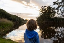Ein kleiner Junge steht in der Abenddämmerung an einem Fluss, Himmelsspiegelungen im flachen, ruhigen Wasser — Stockfoto