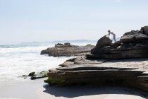 Подросток карабкается по гладким плоским скалам над песчаным пляжем с волнами, ломающимися на берегу. — стоковое фото