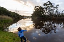 Un ragazzo in piedi vicino a un fiume al crepuscolo, riflessi del cielo nell'acqua piatta e calma — Foto stock