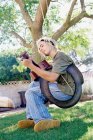 Homem tocando guitarra sentado em um baloiço de pneus em um jardim, cantando. — Fotografia de Stock