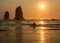 Surfeur marchant hors de la mer avec coucher de soleil derrière la roche dans les vagues sur la plage. — Photo de stock
