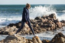 Adolescente équilibrage sur des rochers déchiquetés sur une plage, surf rupture derrière elle. — Photo de stock