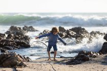 Jovem brincando na areia entre rochas em uma praia, ondas de surf quebrando, — Fotografia de Stock