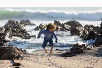 Giovane ragazzo che gioca sulla sabbia tra le rocce su una spiaggia, onde di surf rottura, — Foto stock