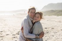 Доросла жінка і її дочка-підліток обіймаються, стоячи на вітряному пляжі — стокове фото