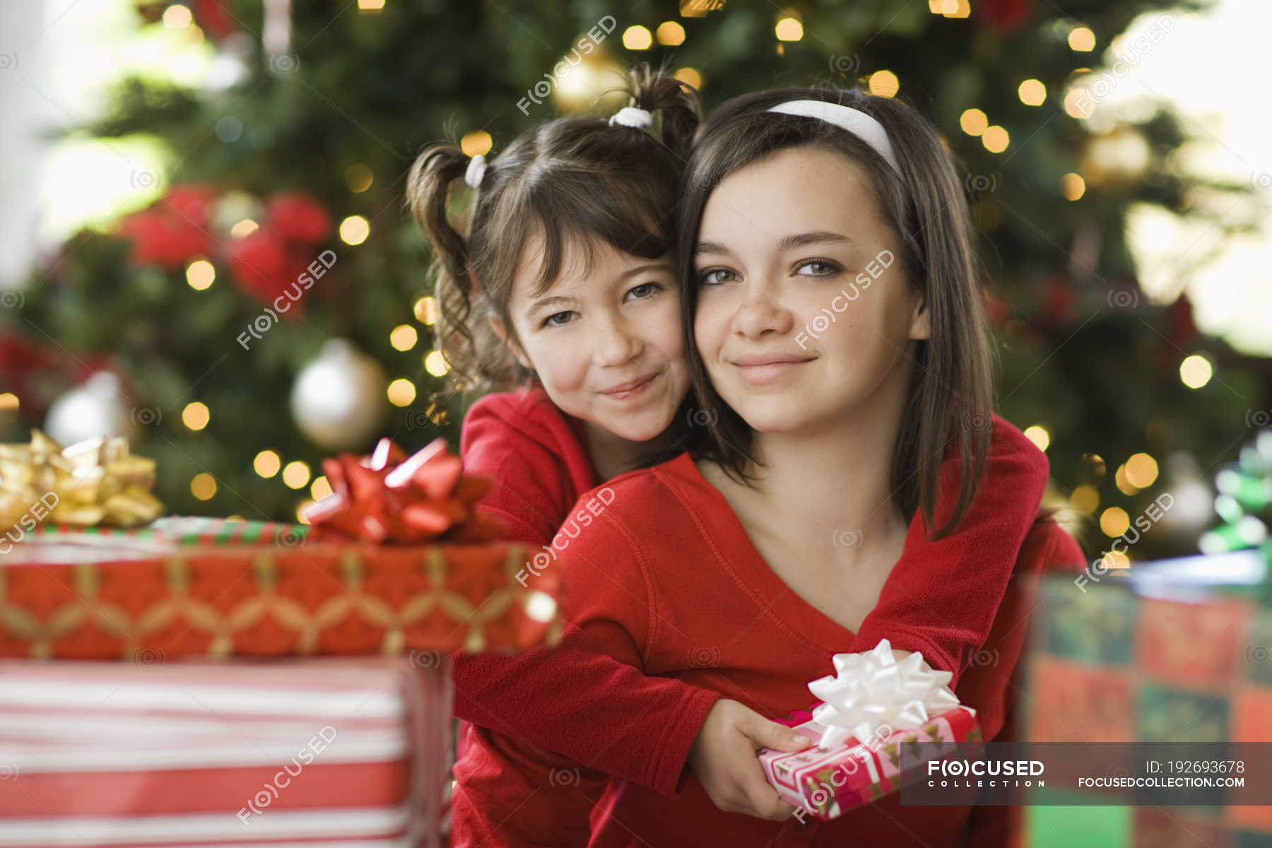 Two girls side by side by Christmas tree surrounded by presents. — kids