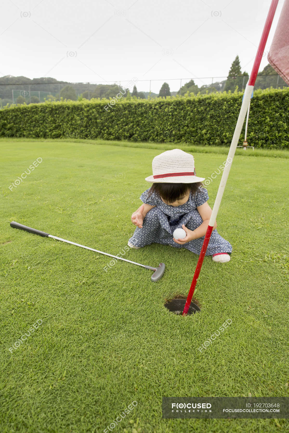 Toddler girl sitting by golf course hole and holding ball. — asian