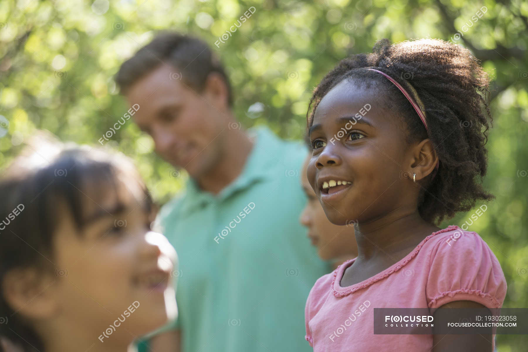 Group of children and mid adult man standing in shade of trees in ...