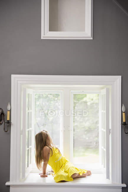 Niña en un vestido amarillo sentado junto a una ventana - foto de stock