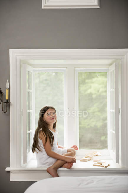 Niña en un vestido blanco sentado junto a una ventana - foto de stock