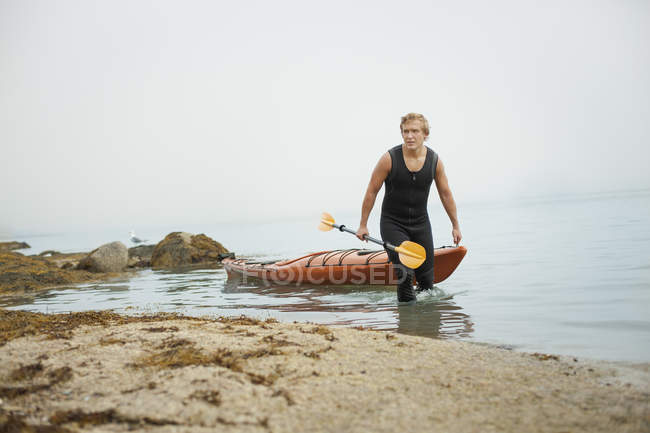 Hombre en traje de neopreno dibujando su kayak - foto de stock