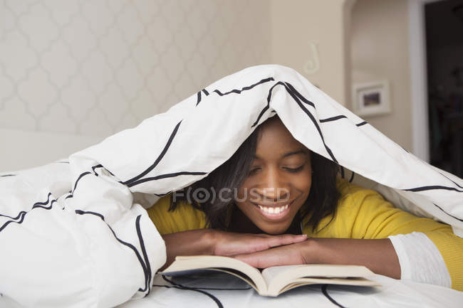 Chica leyendo libro bajo edredón - foto de stock