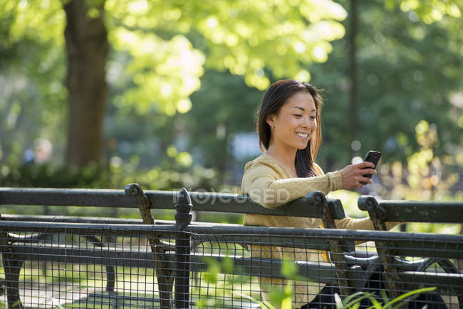 Азиатка проверяет смартфон на скамейке в парке — стоковое фото