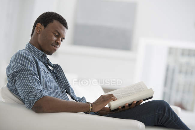 Hombre afroamericano sentado en sillón y leyendo libro . - foto de stock