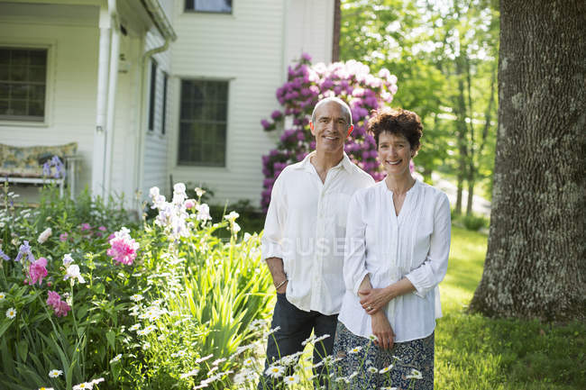 Mature couple in white shirts standing together among flowers in yard. — Stock Photo