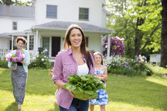 Famiglia di tre generazioni che cammina sul prato portando fiori freschi raccolti, frutta e verdura — Foto stock