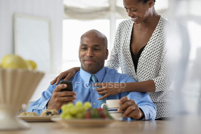 Hombre comprobar teléfono inteligente en la mesa de desayuno con la mujer detrás de enderezar la corbata - foto de stock