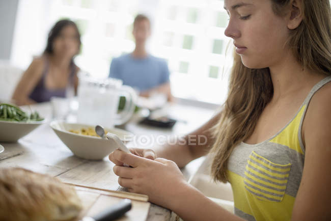 Teenager-Mädchen checkt Smartphone am Esstisch mit Menschen im Hintergrund. — Stockfoto