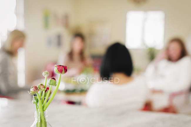 Flores rojas en jarrón con grupo de mujeres alrededor de la mesa en el fondo . - foto de stock