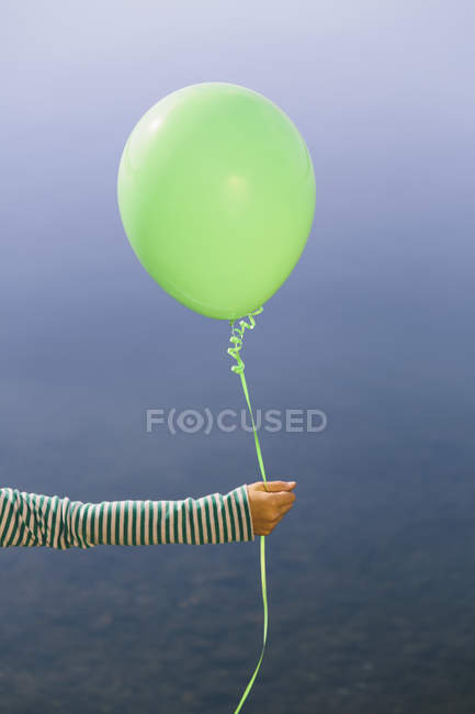 Vista recortada del brazo de la niña sosteniendo globo verde contra el agua del lago - foto de stock