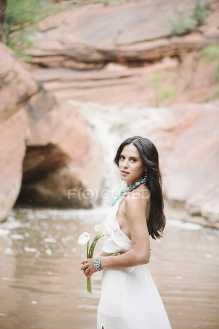 Junge Frau im langen weißen Kleid steht am Fluss und hält Lilien in der Hand. — Stockfoto