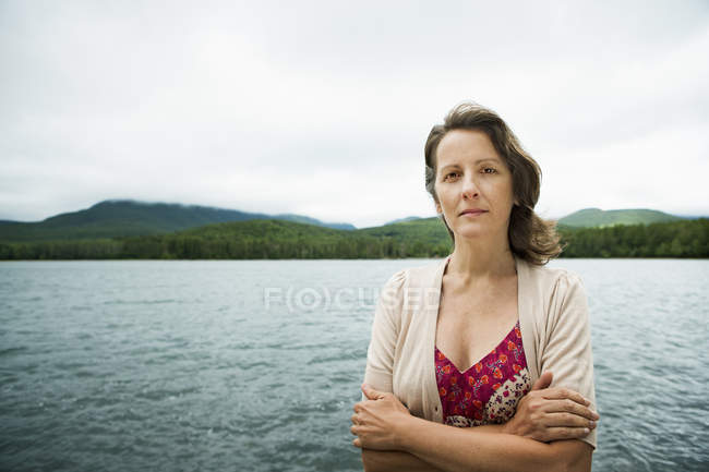 Mulher madura em campo aberto de pé com braços dobrados por lago de montanha . — Fotografia de Stock