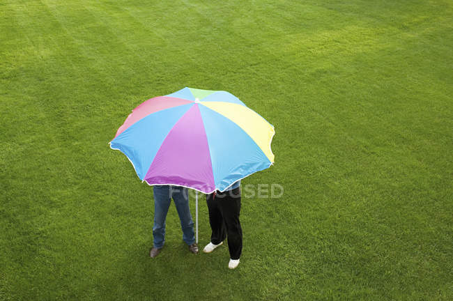Two people under colorful striped umbrella on green lawn. — Stock Photo