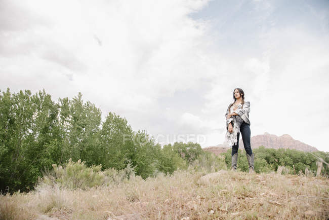 Jeune femme aux longs cheveux bruns debout dans la prairie sous un ciel nuageux . — Photo de stock