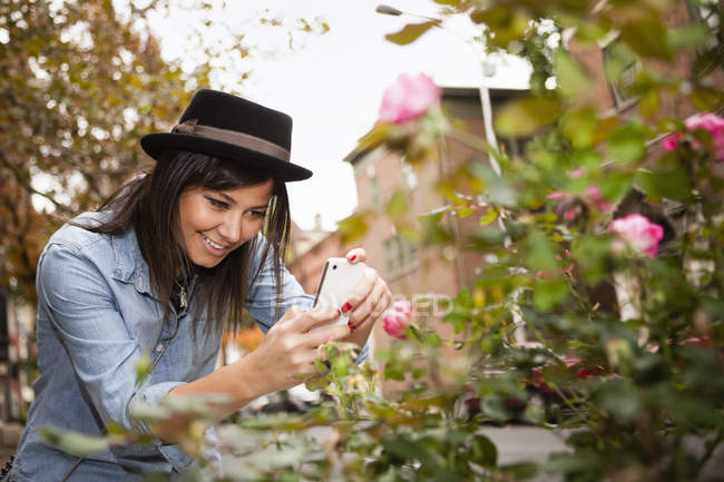 Mujer joven en sombrero trilby tomando fotos de rosas con smartphone . - foto de stock