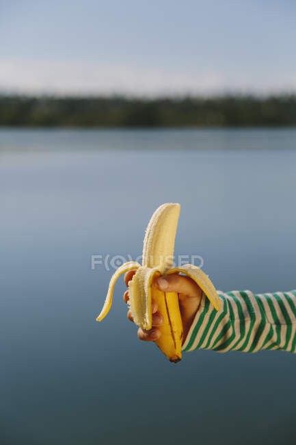 Cropped view of hand of girl holding banana in front of lake — Stock Photo