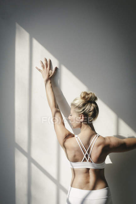 Donna bionda in crop top bianco in piedi davanti alla parete bianca e toccante parete . — Foto stock