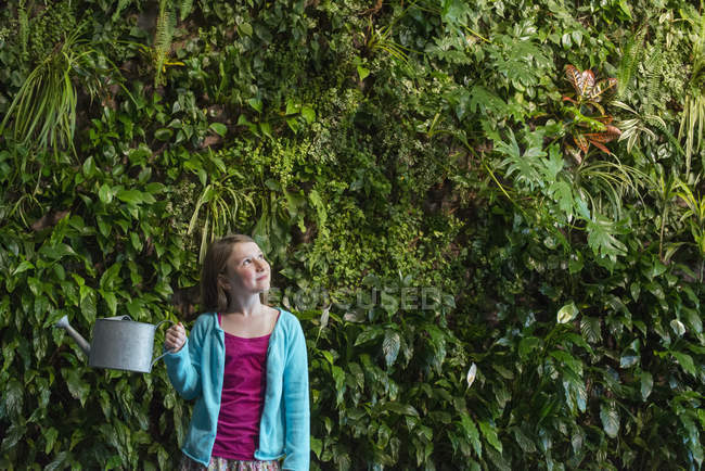 Ragazza in età elementare in piedi di fronte alla parete coperta di felci e piante rampicanti con annaffiatoio
. — Foto stock