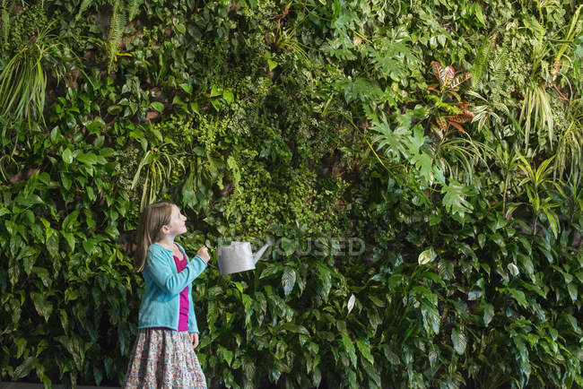 Ragazza in età elementare in piedi di fronte alla parete coperta di felci e piante rampicanti con annaffiatoio . — Foto stock