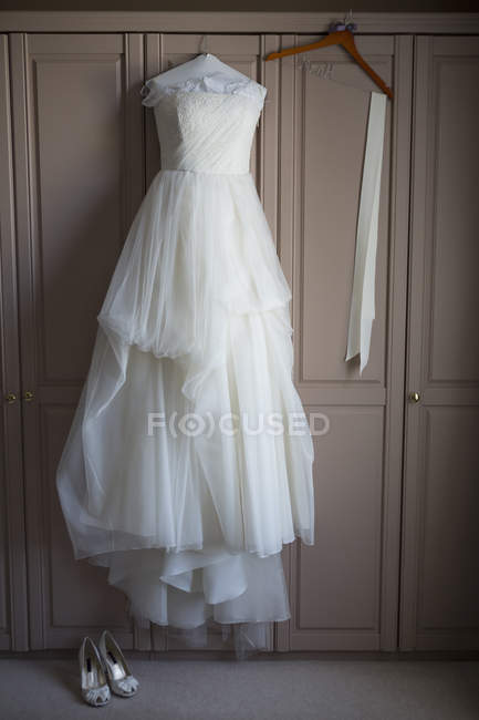 Hochzeitskleid hängt an Schranktür und Hochzeitsschuhe am Boden. — Stockfoto