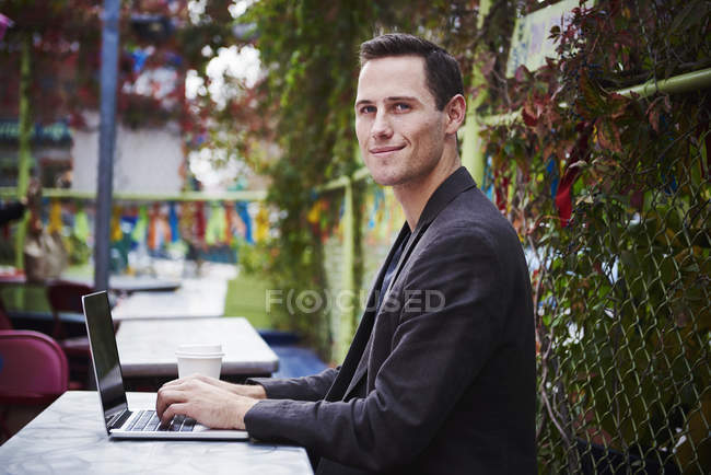 Junger Mann sitzt mit offenem Laptop in der Stadt am Tisch im Freien und blickt in die Kamera. — Stockfoto