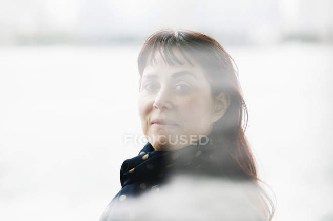 Mature woman with brown hair in scarf looking through window. — Stock Photo
