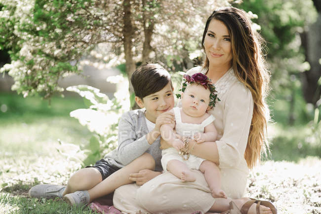 Madre sonriente sentada en el jardín con niño y niña con corona . - foto de stock