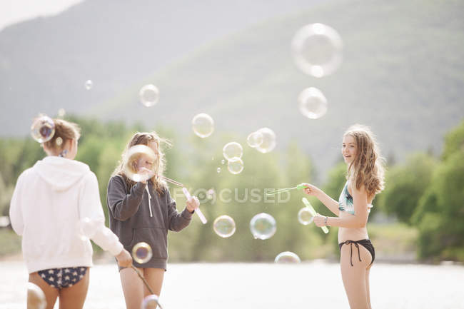 Teenage girls standing by lake surrounded by soap bubbles outdoors. — Stock Photo