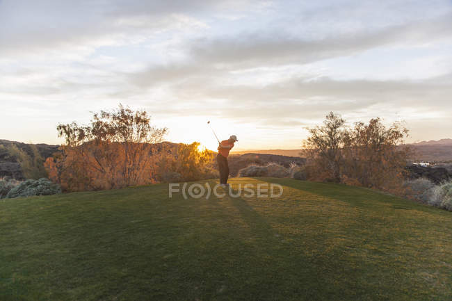 Hombre tomando tiro de la camiseta de golf en el campo de golf en la puesta del sol . - foto de stock