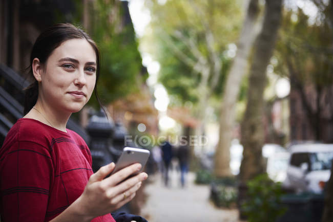 Young woman standing on city street, holding smartphone and looking in camera. — Stock Photo