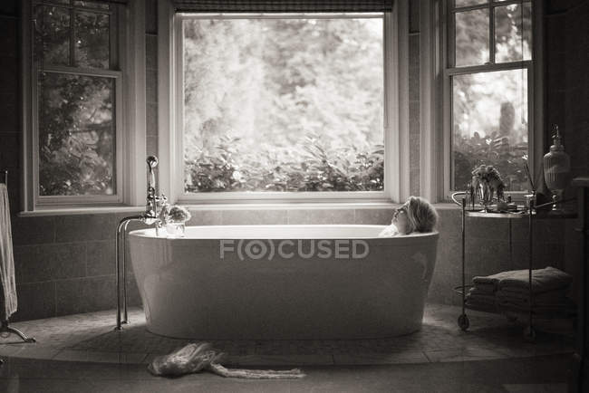 Woman relaxing in free-standing bathtub in bathroom interior with window. — Stock Photo