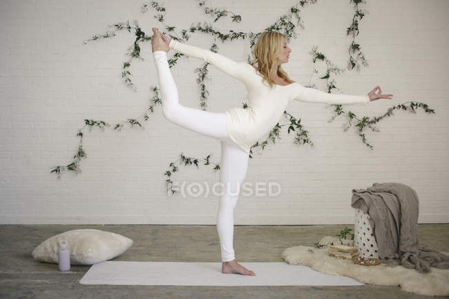 Femme blonde debout sur une jambe avec les bras tendus sur un tapis de yoga blanc . — Photo de stock