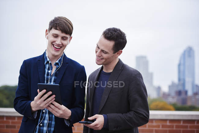Dos hombres jóvenes de pie en la azotea y mirando hacia abajo en la tableta digital juntos . - foto de stock