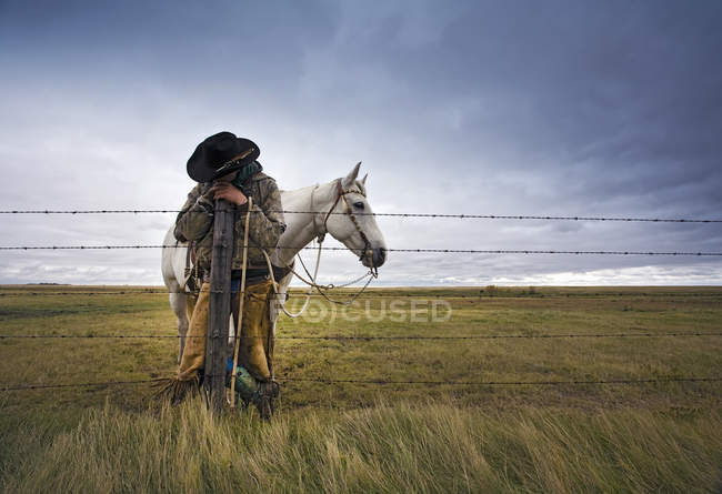 Cowboy apoiando-se no poste de vedação com cavalo cinza atrás no campo . — Fotografia de Stock