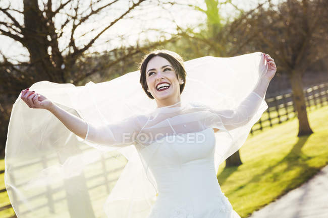 Bride in wedding dress laughing and holding veil outdoors. — Stock Photo