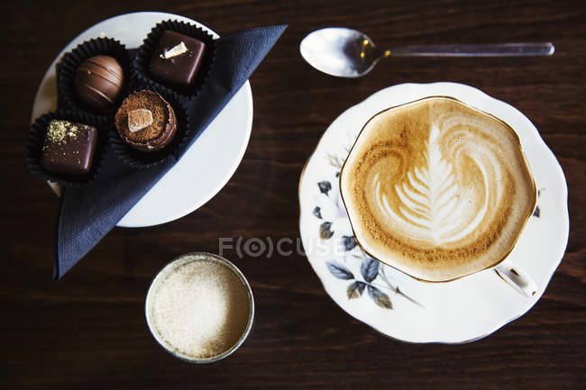 Surtido de caramelos de chocolate hechos a mano en el plato con taza de café con patrón de espuma . - foto de stock