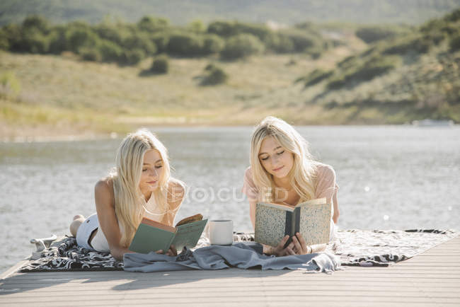 Two blonde teenage girls lying on lake jetty and reading books. — Stock Photo