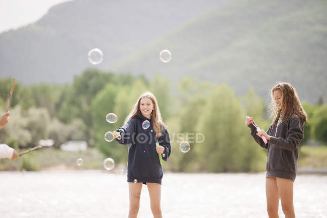 Ragazze adolescenti in piedi vicino al lago circondate da bolle di sapone all'aperto . — Foto stock