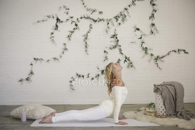 Femme blonde allongée sur un tapis blanc et faisant une pose de yoga pour chien orientée vers le haut . — Photo de stock